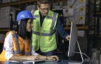  A female worker wearing a blue safety helmet and orange reflective vest uses a digital tablet while discussing information with a male colleague in a neon green reflective vest. They are collaborating in a warehouse environment, surrounded by stacked boxes and logistics equipment
       -  (crédito:  Getty Images)